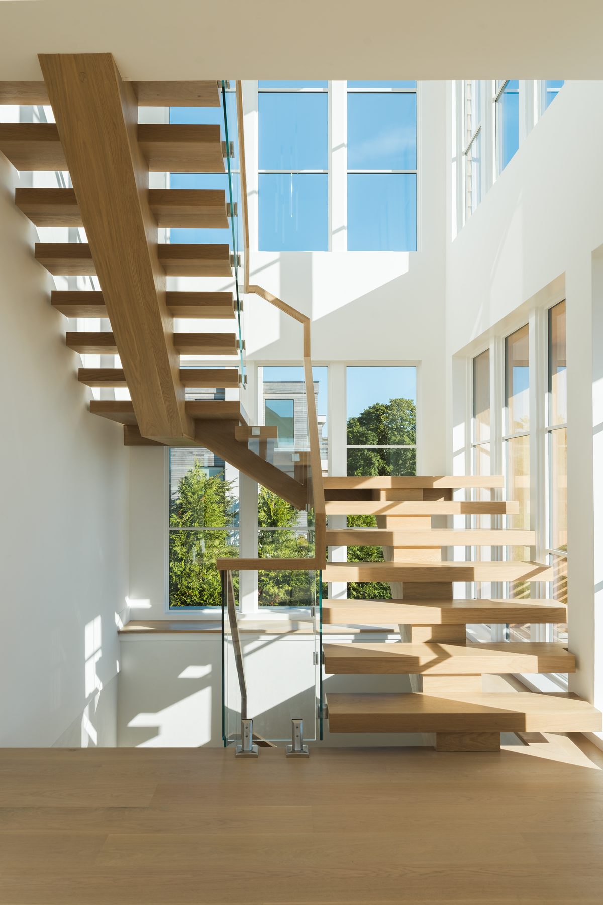 A floating light-oak staircase with glass railings rising through a sunlit, double-height stairwell lined with tall windows