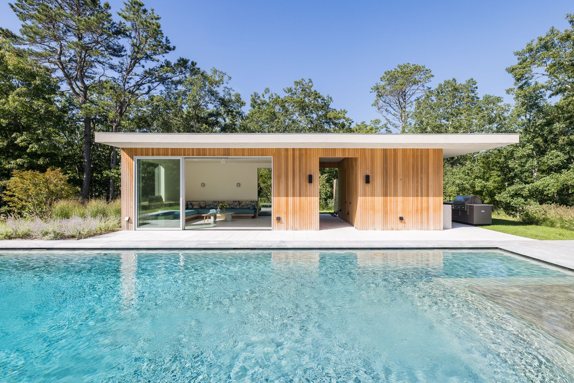 Flat-roofed pool house with its sliders open, seen straight across the pool